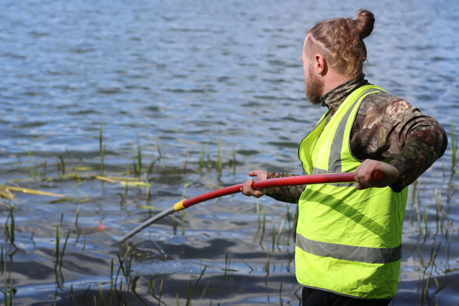 An angler in hi-vis working the margins of a still water with a long-handled tool.
