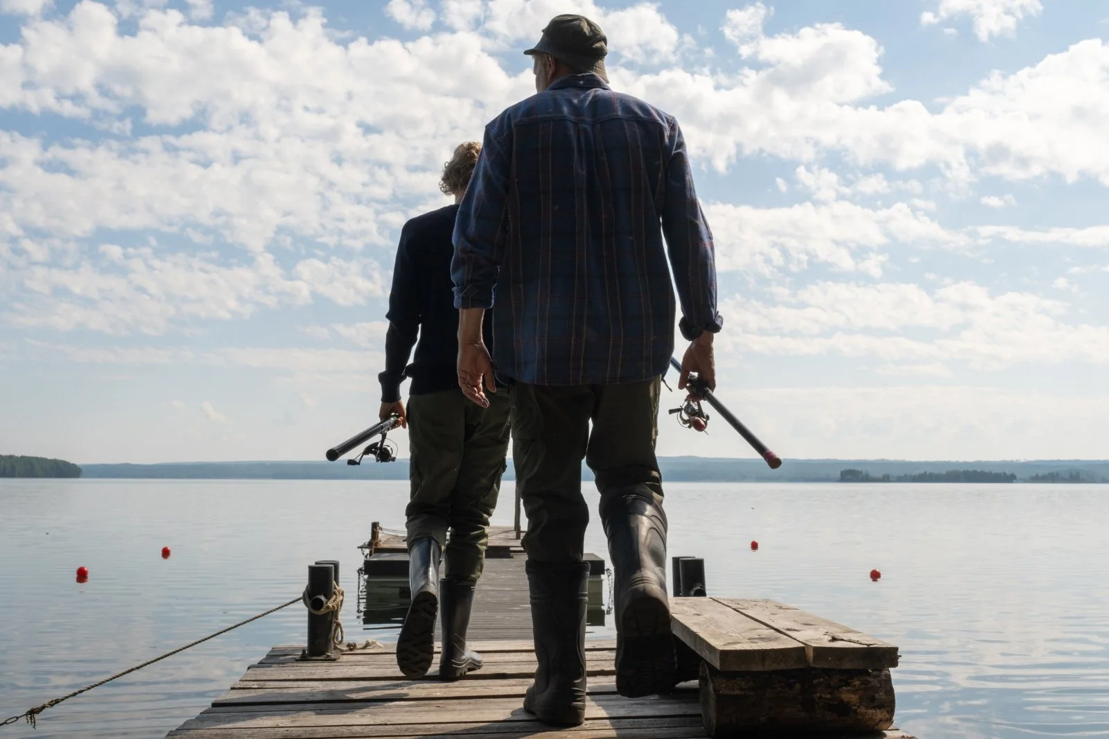 Two anglers walking together down a wooden jetty toward open water, rods in hand, backs to camera. An older man in a checked shirt and a younger figure ahead, under a wide cloudy sky.